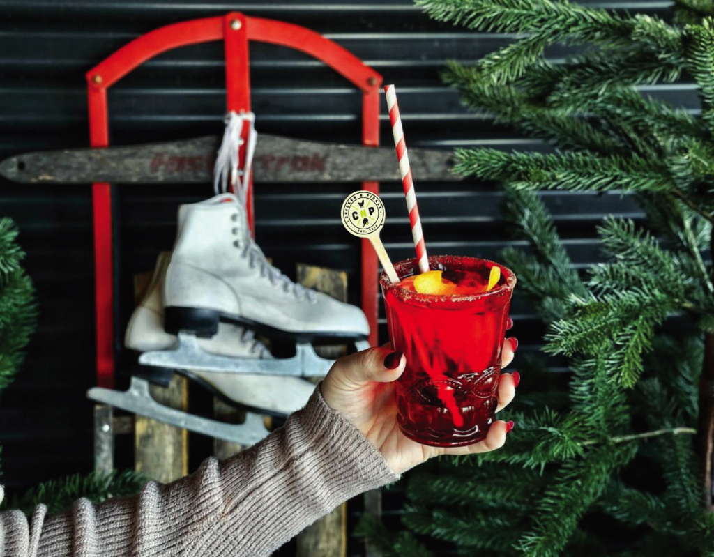 A hand holds a vibrant red cocktail garnished with orange slices and a striped straw. In the background, there is a vintage ice skate on a red sled, surrounded by green pine branches.